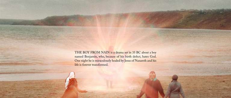 Scene from screenplay 'The Boy from Nain,' a drama set in 35 BC written by Cy Young. The film explores themes of faith and transformation, following Benjamin's miraculous healing by Jesus of Nazareth. The scene shows three people on a beach at sunset facing a heavenly, illuminated figure. The people have open arms and posture to indicate worship and awe. The scene is a tranquil atmosphere and vibrant colors. An overlay of text says, 